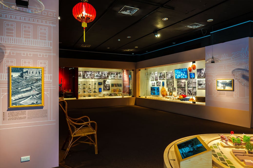 展區實景：左側牆面掛有黑白攝影作品與紅燈籠，前方放置復古藤椅；後方玻璃展櫃陳列老照片與文物，右下角為街道模型與觸控螢幕。