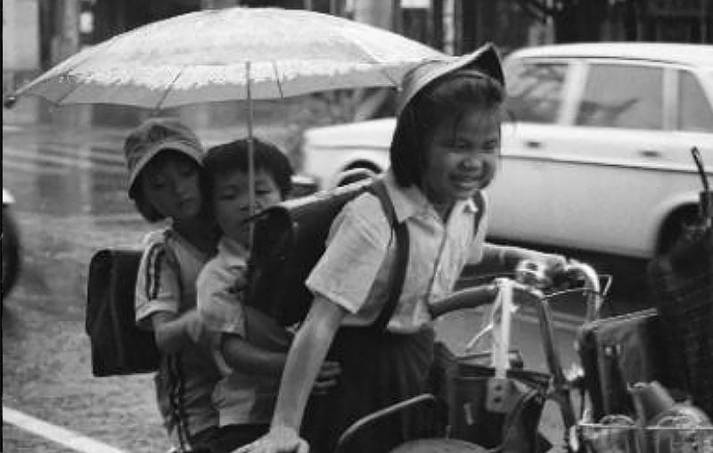 雨中三位穿校服的小學生共乘一輛腳踏車，姐姐奮力踩車、面帶笑容，後座弟妹共撐一把小傘。
