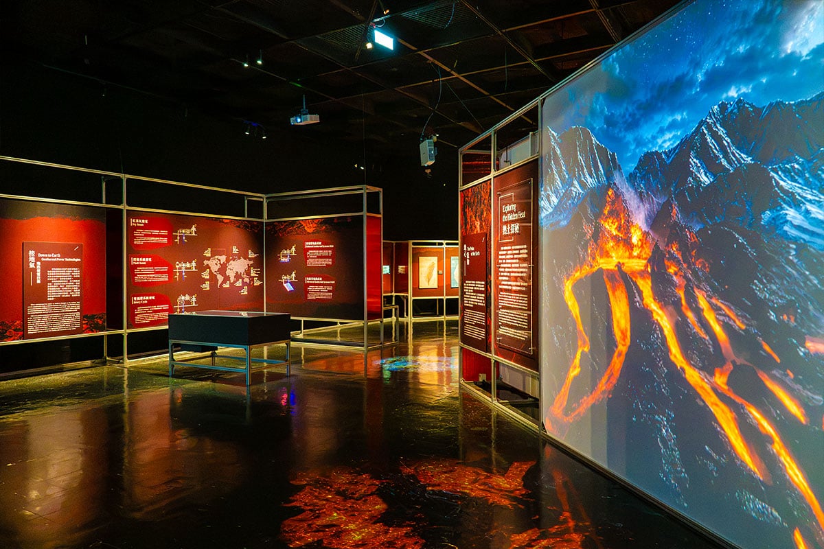 『熱土探秘』展區實景：右側牆面有火山噴發與岩漿流動的大型投影，暗色展場內設有紅色主題展板與展示台。