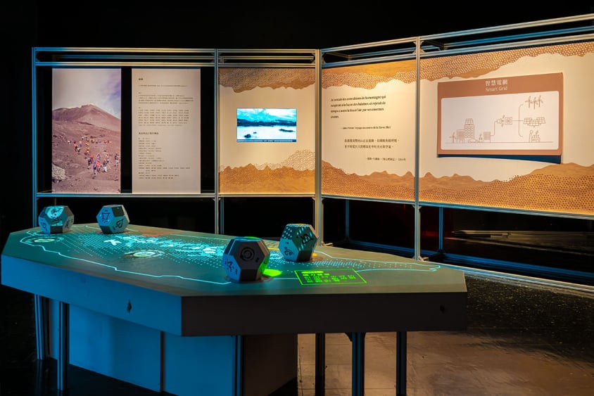 『永續同行』展區實景：前景為發光的藍色互動展台與骰子裝置，背景為金屬框架說明展板與螢幕